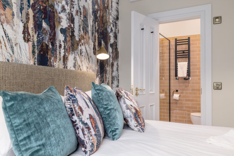 Double bed with teal cushions beside an open bathroom door