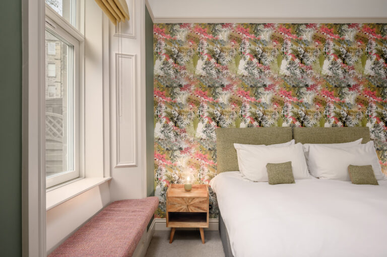 Twin beds beneath floral wallpaper beside a tall window