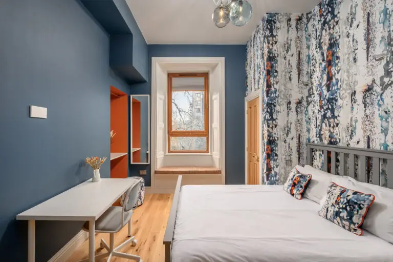 Double bed beside a desk and tall window in a blue studio room