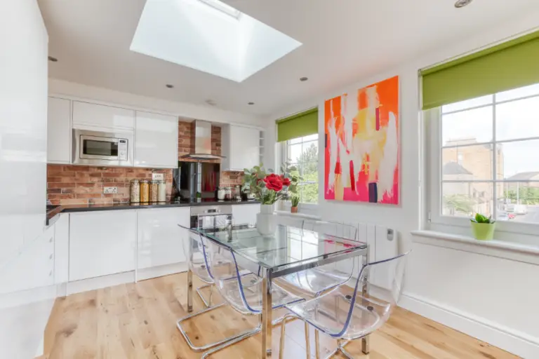 Glass dining table beside a white kitchen with exposed brick walls